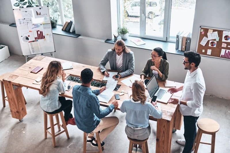 Six colleagues around a tall wooden table, reviewing printed charts and a laptop together.