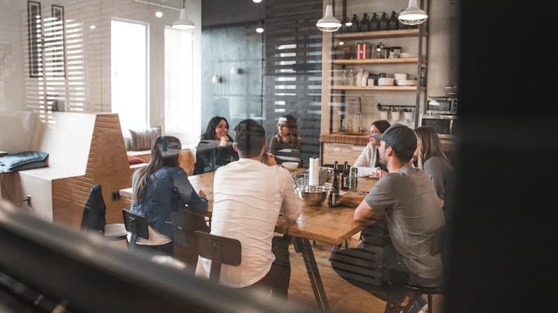 A team sharing a long table over lunch in an open, daylit office kitchen.
